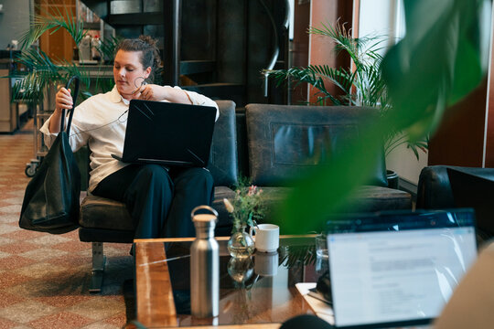 Female entrepreneur holding her bag while sitting with open laptop on sofa in coworking space lounge