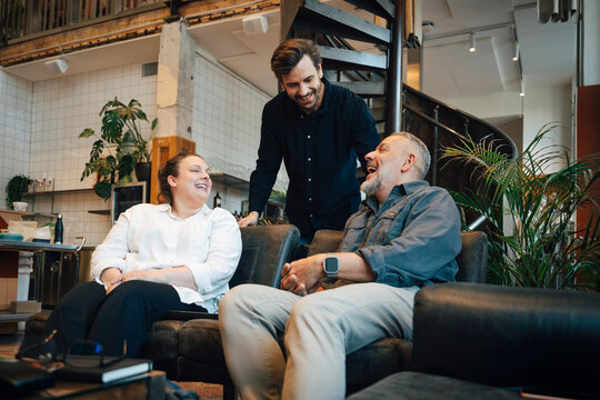Happy coworkers laughing together while sitting on couch in modern office lounge