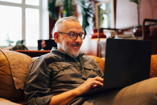 Smiling male entrepreneur working on laptop while sitting comfortable on sofa in coworking space lounge