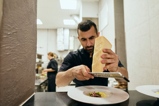 Focused male chef grating cheese over dish at kitchen pass window