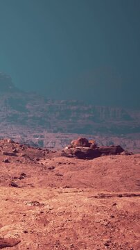 A rocky surface stretches across the landscape. The ground is covered with red soil and various stones. A distant horizon reveals more rugged terrain under a sky with a strange hue.