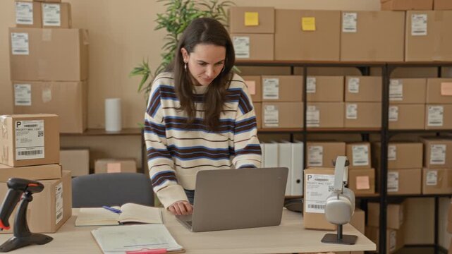 Woman at laptop smiling while handling parcels and scanning packages in a building; small business optimism.