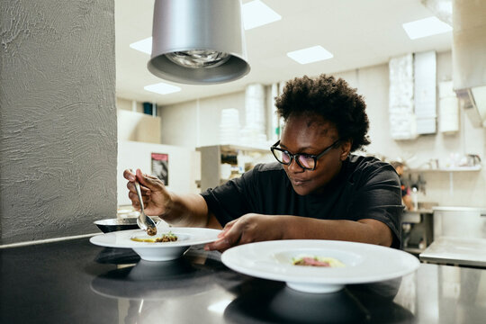 Dedicated female chef using spoon to garnish gourmet dish at kitchen pass window