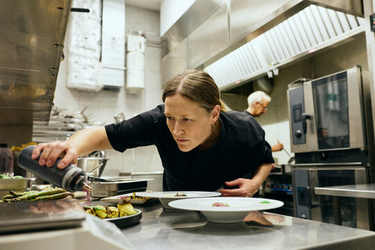 Focused female chef using plastic bottle to add sauce to plates in busy commercial kitchen