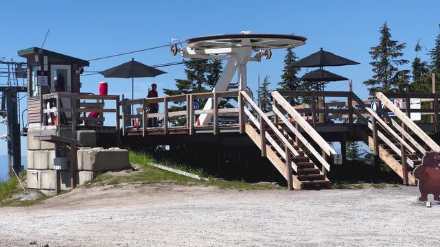 The top station of the Peak Chairlift at Grouse Mountain, its bullwheel, wooden viewing deck, patio umbrellas and stairs under a blue summer sky in North Vancouver