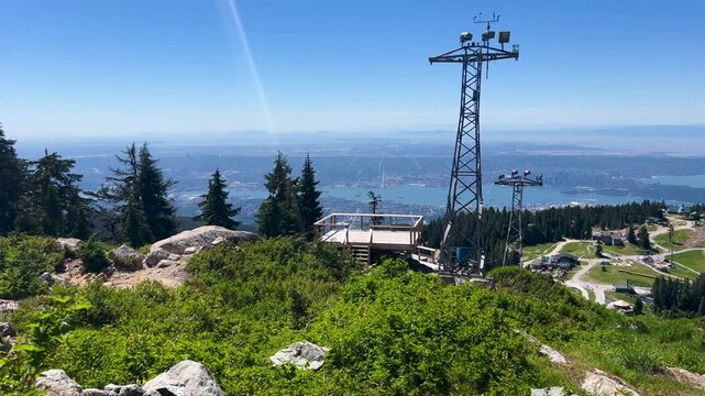 A communications tower and an observation deck rise at the Grouse Mountain summit, subalpine shrubs and the city and Burrard Inlet far below in summer haze