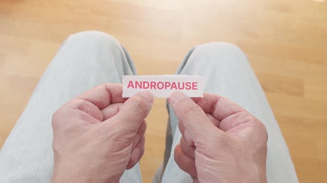 A first-person view of a man holding a torn piece of paper with the word Andropause printed on it, symbolizing male aging and hormonal changes. Close up.