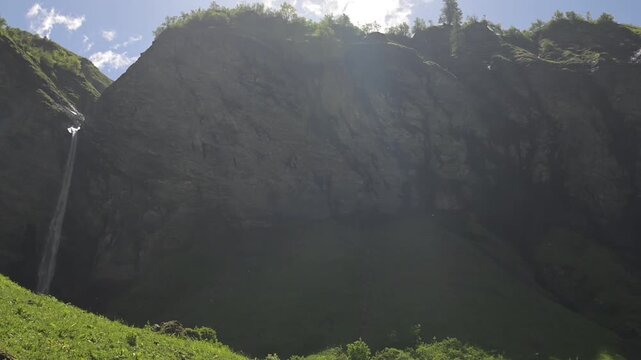 Summer landscape of a waterfall flowing down steep Swiss cliffs with sun rays and green vegetation.
