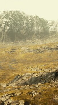 Scenic view shows mountains in the background with peaks covered in snow. Grasslands stretch in the foreground with rocky terrain. The sky appears bright and clear above the land.