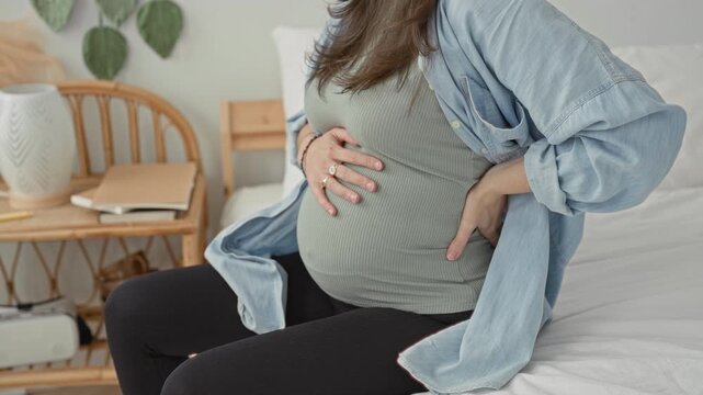 Pregnant woman clutching baby bump, hand on lower back for backache, sitting on bed in building; concern support.