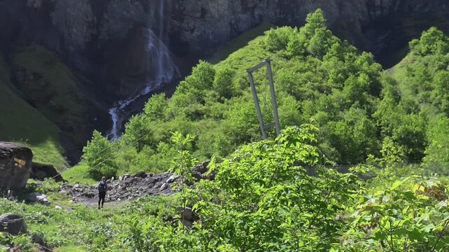 Arena Batoni, Switzerland - 16.06.2026: Hikers walk along rocky mountain path with green hillsides and a waterfall in the background.