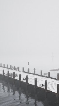 A wooden pier extends over calm water. Fog fills the area, making it hard to see. There are few visible objects far away, creating a mysterious scene in the morning.