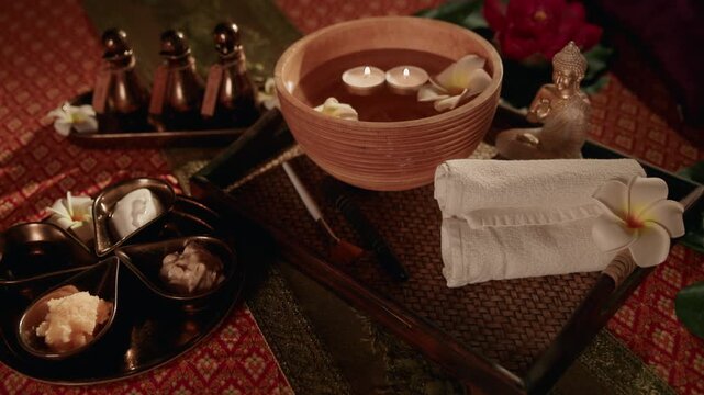 Slow motion close up of spa bowl with floating candles towel and frangipani on woven tray in dim Thai massage room.