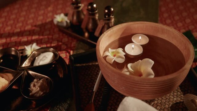 Wooden bowl with floating candles and plumeria flowers on spa treatment table with gentle pan tracking, Thai relax Oil.