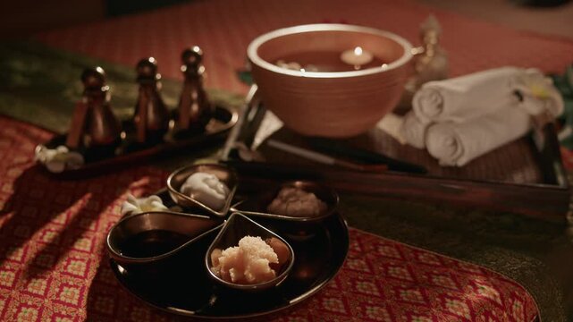 Thai spa setup with ceramic bowls oils and rolled towels on patterned table in warm candlelit treatment room.