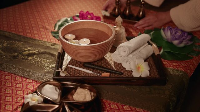 Top view spa tray with wooden bowl candles and rolled towels on massage table as therapist reaches in for arrangement, gentle lateral pan.
