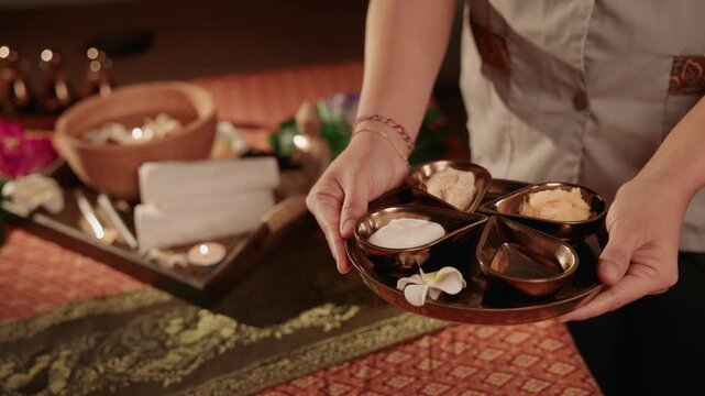 Handheld tracking shot of Thai spa therapist presenting bowls of oils and creams on wooden tray in treatment room, slight pan.