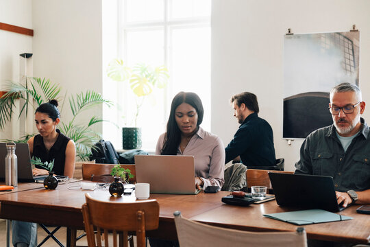 Diverse freelancers managing their tasks on laptops while working at shared open-plan workspace table