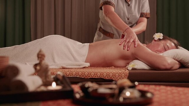 Slow motion woman receiving back Thai massage from masseuse on spa treatment table in dim candlelit room.