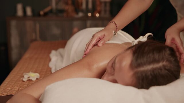 Slow motion Thai masseuse massaging woman on spa massage table in dim treatment room, warm light.