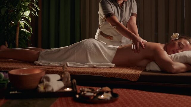 Slow motion masseuse massaging female client on spa treatment table in candlelit wellness room with plants.