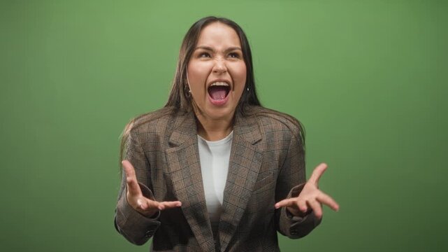 Woman in plaid blazer and white tee with raised hands and open mouth gesturing emphatically against a studio green screen, hoop earrings visible; frustration.
