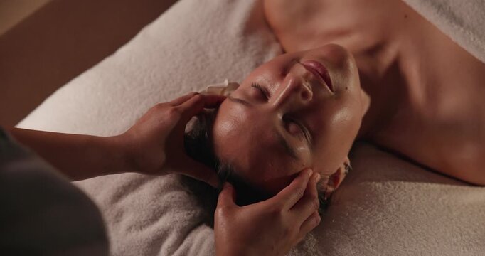 Tracking shot woman receiving head and facial massage on spa treatment table in warm indoor room slow motion handheld close.