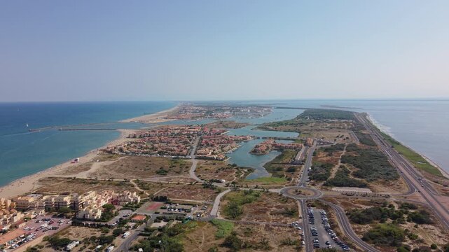 Drone scenic tracking shot of Port Leucate coastal resort town, showing Mediterranean beaches, holiday apartments, inner marinas, and the highway infrastructure in Occitanie, France.