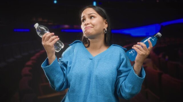 Woman holding two bottles and shrugging inside a dim theater building with rows of seats and blue accent lighting; indecision choice refreshment.