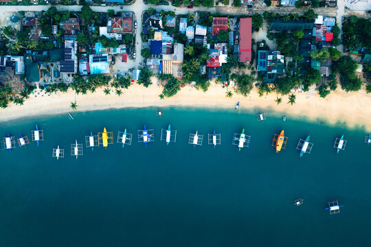 Aerial view of Port Barton beach with colorful outrigger boats anchored in turquoise water, lined with tropical trees and coastal buildings in San Vicente, Mimaropa, Philippines.