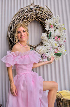 Beautiful mid adult woman in pink evening dress posing near large decorative white flower wreath.