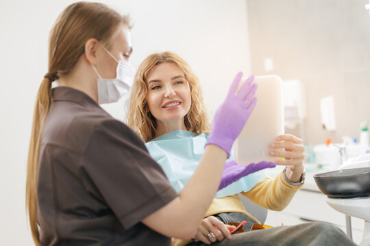 Young caucasian female dentist and patient examining smile in modern dental clinic after correction restoration of chipped front broken tooth of woman