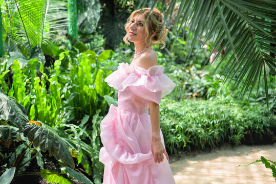 Beautiful romantic woman in elegant pink tulle dress dreaming in tropical greenhouse. Sensual blonde girl posing in botanical garden orangerie surrounded by palm trees.