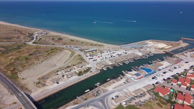 Drone flight over the coastal road bridge and oyster market channel, revealing the sandy beach, blue Mediterranean sea with sailing boats, and tourist infrastructure in Leucate, France.