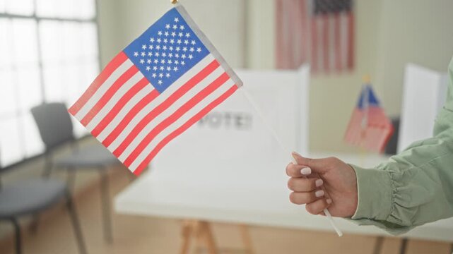 Woman holding small american flag with visible hand at a pollingstation voting booth; pride civic duty.