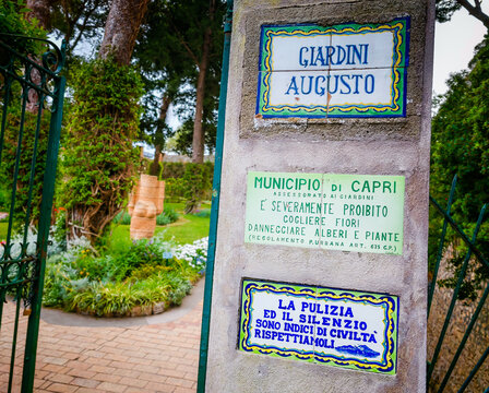 07 02 2026 - Capri, Italy. Entrance to Giardini Augusto, featuring a signboard display in foreground with a concrete and brick pathway adorned by vibrant flowers and trees, set against a lush green