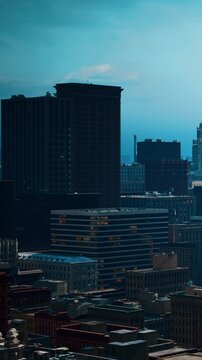Bright sunlight shines on tall buildings in a city. The view shows various structures with different designs. The skyline has a mix of old and modern architecture.