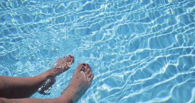Slow motion footage of a pair of woman&rsquo;s feet gently splashing in a clear blue swimming pool with shimmering water reflections creating relaxing holiday scenes at a luxury resort during the summer