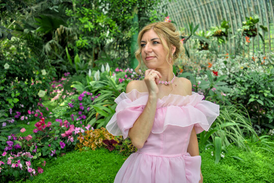 Waist up portrait of a thoughtful beautiful caucasian woman in an aesthetic pastel pink gown decorated with ruffles, posing against dense green foliage in a botanical garden.