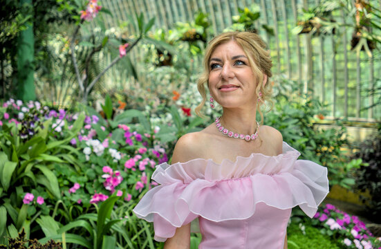 Portrait of a happy charming caucasian female model wearing elegant off shoulder pink ruffle dress posing in a lush green tropical greenhouse conservatory.