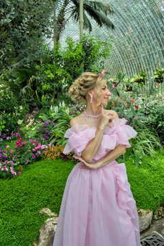Full length portrait of a sophisticated graceful caucasian woman wearing an aesthetic pastel pink dress inside a spectacular glass dome orangery surrounded by exotic plants and palm trees.