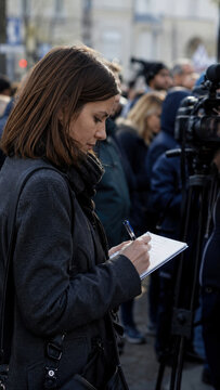 Focused female journalist stands in crowded outdoor setting, diligently taking notes in her notebook while covering public news event with cameras in background.
