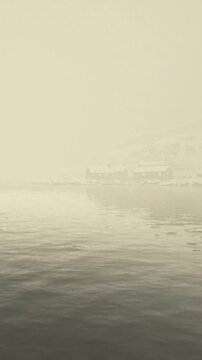 Fog hangs over the water, making the scene appear soft and blurred. Buildings can be seen along the shore, shrouded in mist on a cloudy day. Waves gently ripple on the surface.