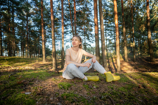 Smiling tranquil woman relaxing in autumn forest alone, sitting on meadow under warm sunlight, female nature lover observing wild woodlands and calming mind, recreational leisure trip in wilderness.