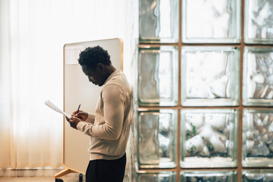 Side view of businessman writing on documents while leaning on glass brick wall