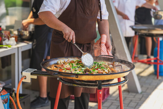 Chef cooks paella in pan with seafood, rice, herbs, steam during outdoor catering. Paella connects Spanish cuisine, street food, festival dining, restaurant service, gastronomy