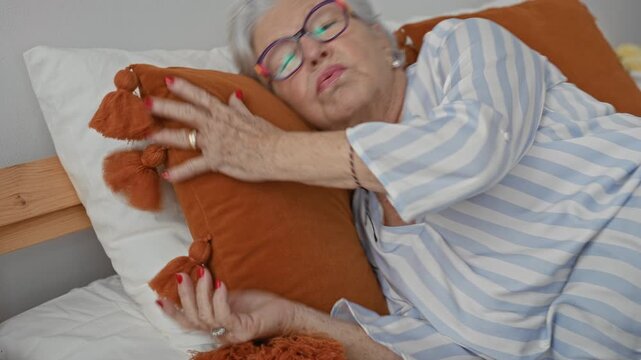 Woman hugging an orange cushion with bare hands while lying on a bed in a building, wearing striped pajamas and glasses, head resting on pillow; comfort.