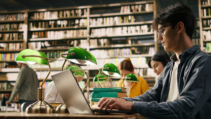 Young student typing on laptop in quiet library study area near, bookshelves with study session. © DC Studio