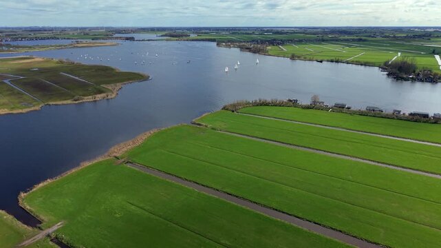 High-angle aerial drone panoramic view of traditional Dutch polder landscape with structured green agricultural fields, drainage canals, and numerous white sailboats navigating a vast open lake