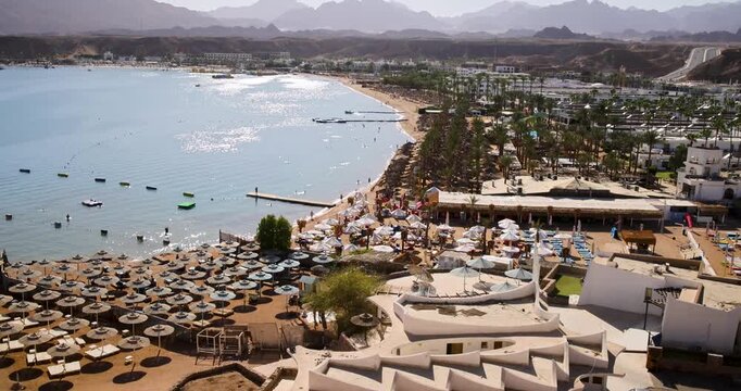 Slow motion footage aerial view of Naama Bay in Sharm El Sheikh Egypt showing a busy beachfront with parasols sun loungers resort hotels coastal attractions and visitors enjoying the Red Sea shoreline